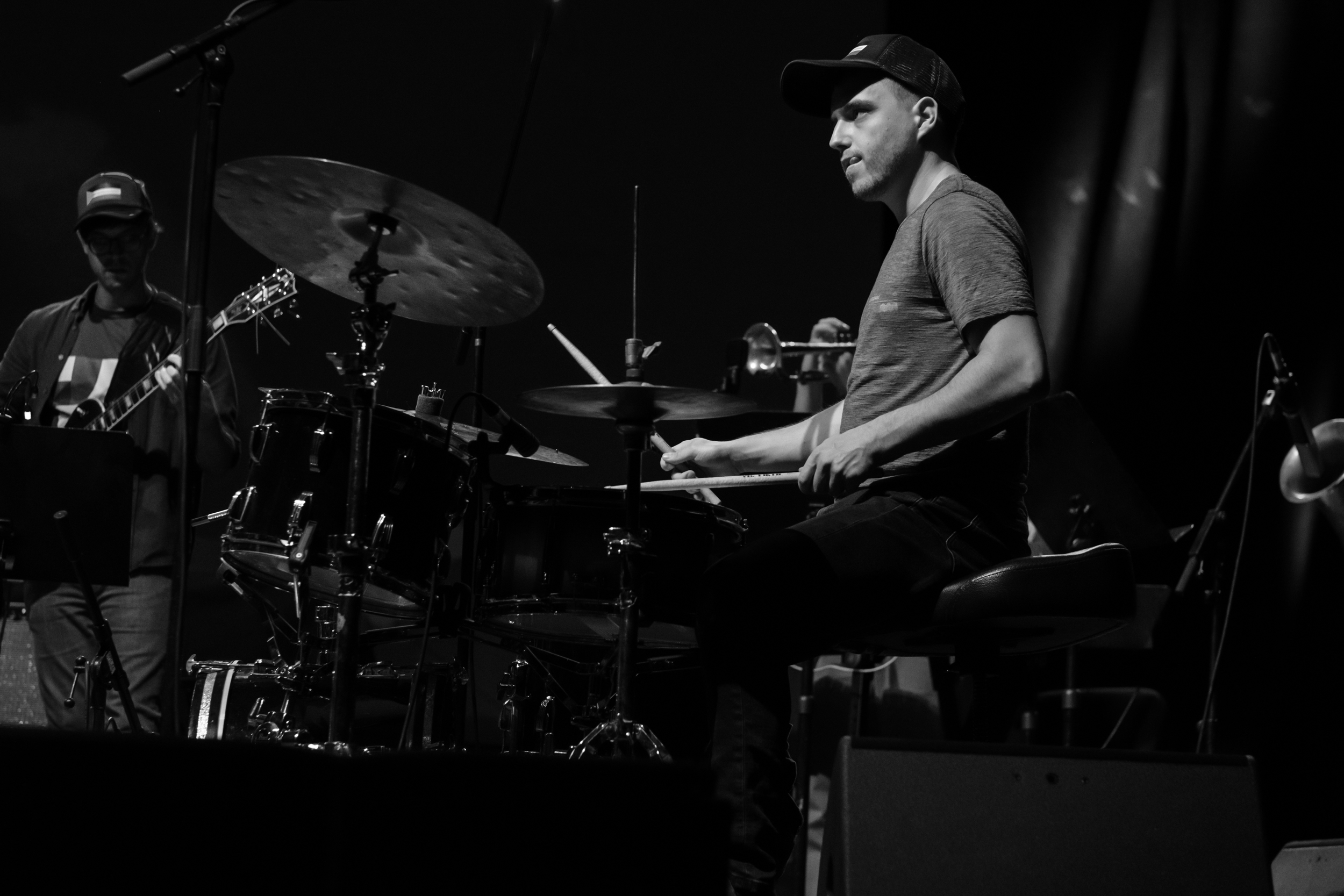 A drummer plays at a drum kit, with another musician partly visible to the left.