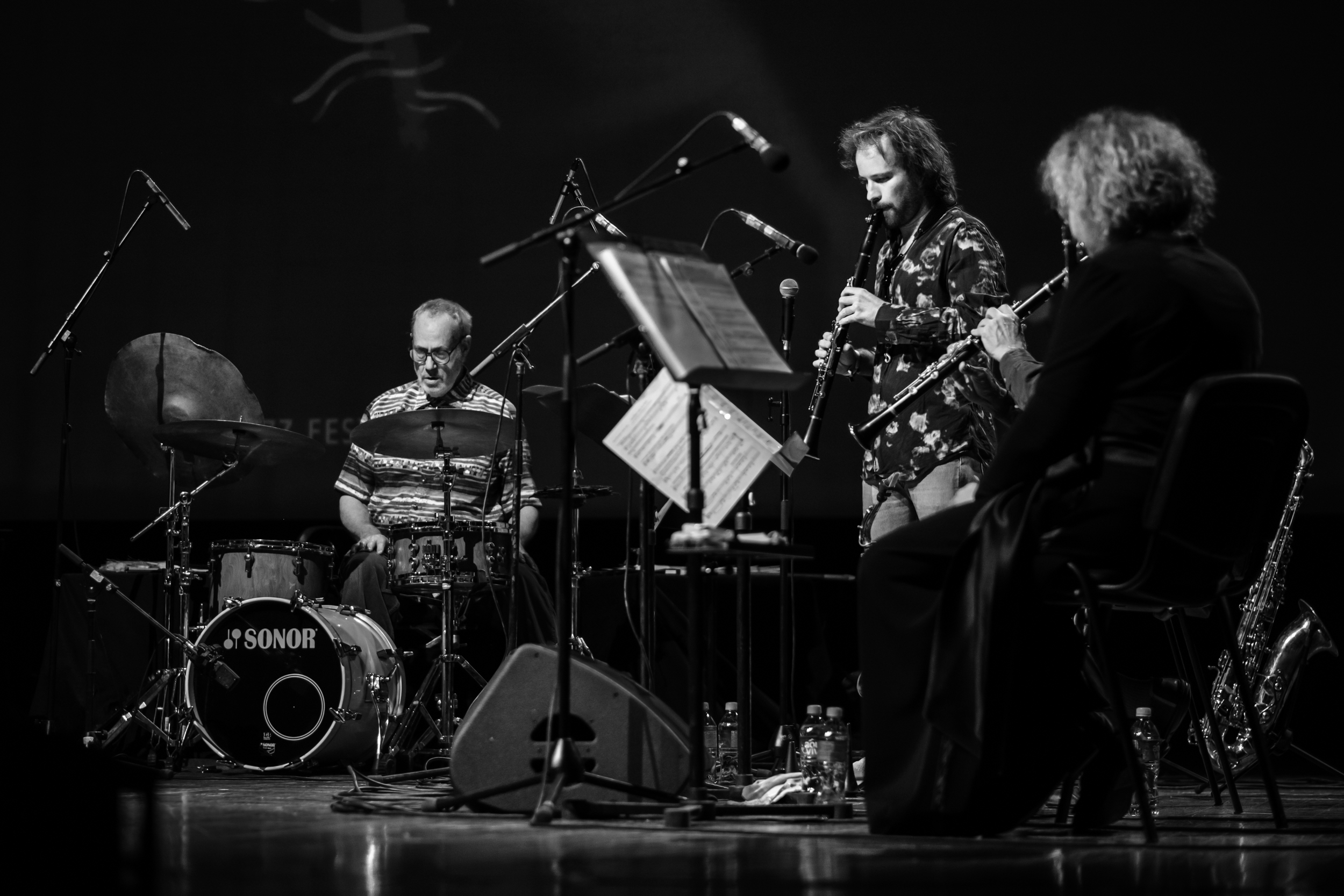 A saxophonist performs near a drum kit and seated fellow musicians.