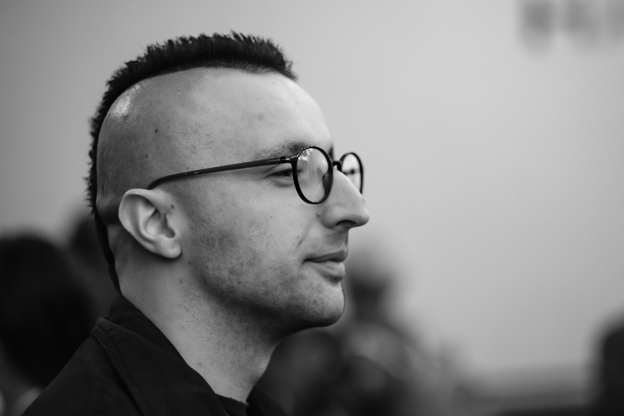 A man wearing round glasses is shown in side profile in a crowded foyer.