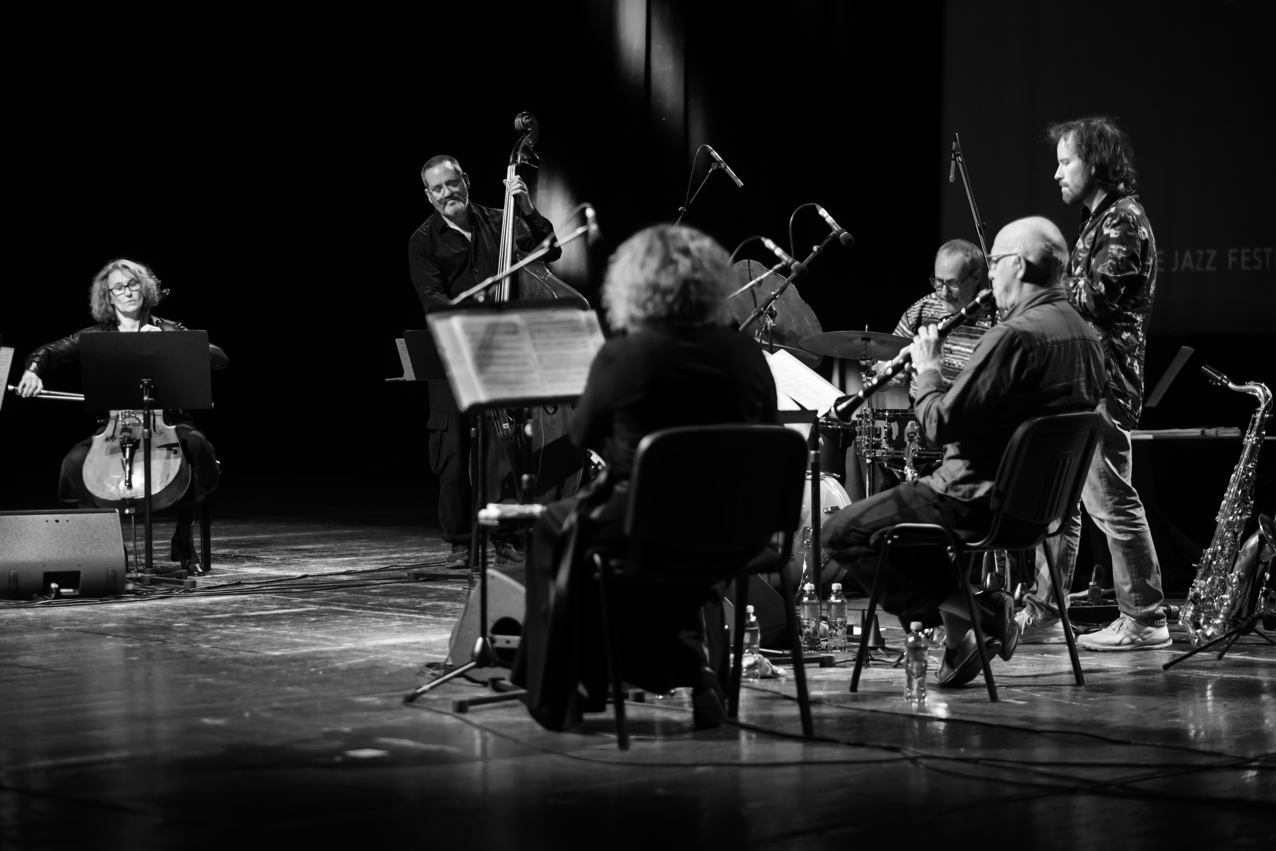 A large ensemble performs, with a double bass player and seated instrumentalists.