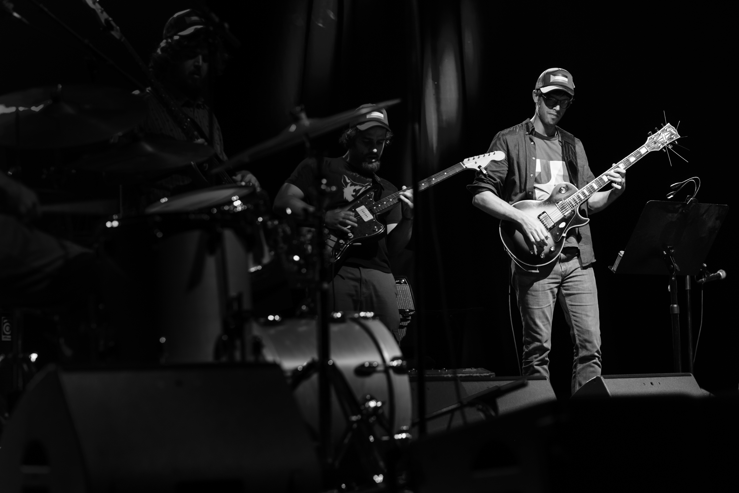 A guitarist plays in a pool of stage light, with other musicians in shadow.